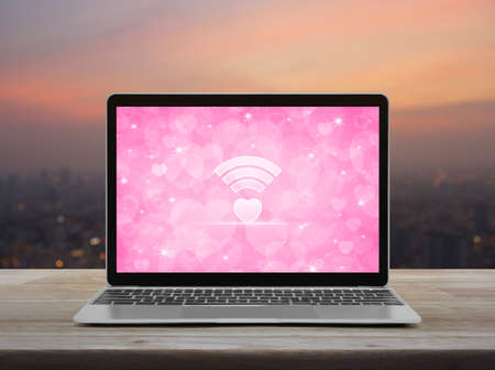 Heart love wifi flat icon modern laptop computer with pink screen on wooden table over blur of cityscape on warm light sundown, Internet online love connection, Valentines day conceptの写真素材