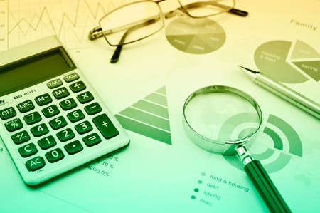 Magnifying glass, calculator, pen, and glasses on business budget graph, green and yellow tone,...