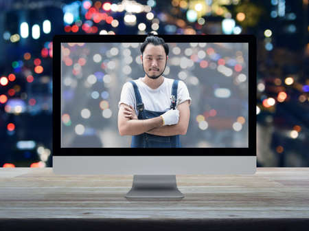 Asian mechanic car engine in uniform with tools on desktop modern computer monitor screen on wooden table over blur colorful night light traffic jam road in city, Business repair car service online conceptの写真素材