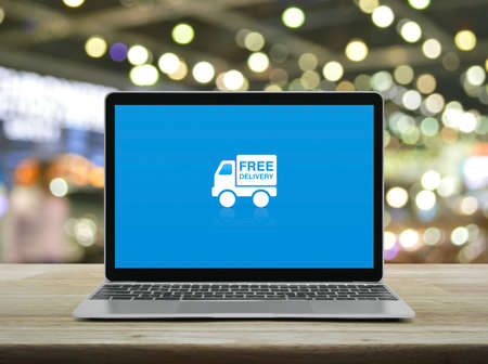 Free delivery truck flat icon with modern laptop computer on wooden table over blur light and shadow of shopping mall, Business transportation online conceptの写真素材