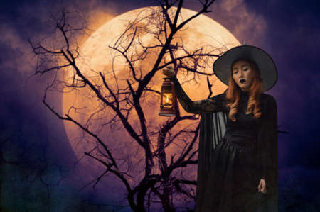 Halloween witch holding ancient lamp standing over dead tree, full moon and spooky cloudy sky, Halloween mystery conceptの写真素材