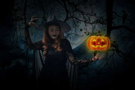 Halloween witch with pumpkin monster head standing over dead tree, crow, birds, full moon and spooky cloudy sky, Halloween mystery conceptの写真素材