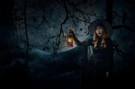 Halloween witch holding ancient lamp standing over dead tree, crow, birds, full moon and spooky cloudy sky, Halloween mystery conceptの写真素材
