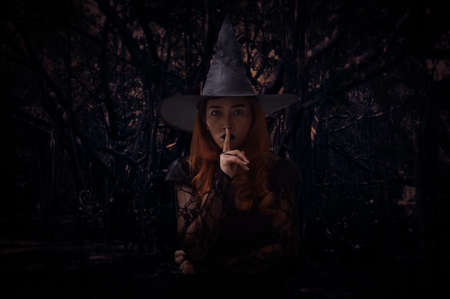 Halloween witch showing silence sign with finger over lips standing over spooky dark forest with tree, leaves and vine, Halloween mystery conceptの写真素材