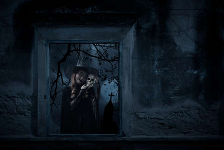 Halloween witch holding a skull standing in old damaged window with wall over cross, church, birds, dead tree and spooky cloudy sky, Halloween mystery conceptの写真素材