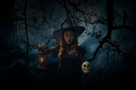 Halloween witch holding ancient lamp and skull standing over dead tree, crow, birds, full moon and...