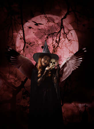 Halloween witch with wings holding a skull standing over cross, church, crow, birds, dead tree, full moon and cloudy spooky sky, Halloween mystery conceptの写真素材