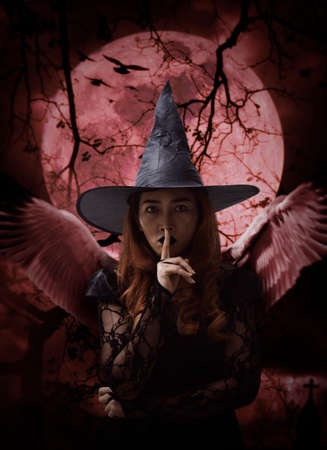 Halloween witch with wings showing silence sign with finger over lips standing over cross, church,...