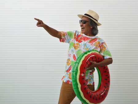 Laughing elderly traveler asian man wearing summer shirt, straw hat and sunglasses holding inflatable ring, pointing finger away, standing over white wall background, Business summer holiday conceptの写真素材