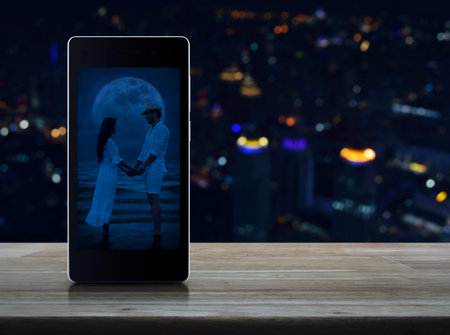 Romantic loving couple standing and holding hands together on modern smart mobile phone screen on wooden table over blur colorful night light city tower and skyscraper, Business internet dating online, Valentines day conceptの写真素材