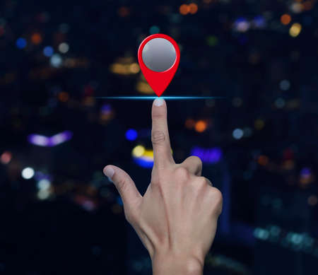 Hand pressing map pin point location button over blur colorful night light modern city tower and skyscraper, Map pointer navigation conceptの写真素材