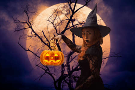 Halloween witch with pumpkin monster head over dead tree, full moon and spooky cloudy sky, Halloween mystery conceptの写真素材