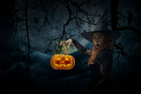 Halloween witch with pumpkin monster head standing over dead tree, crow, birds, full moon and spooky cloudy sky, Halloween mystery conceptの写真素材