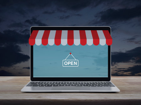 Modern laptop computer with online shopping store graphic and open sign on wooden table over sunset sky, Business internet shop online conceptの写真素材