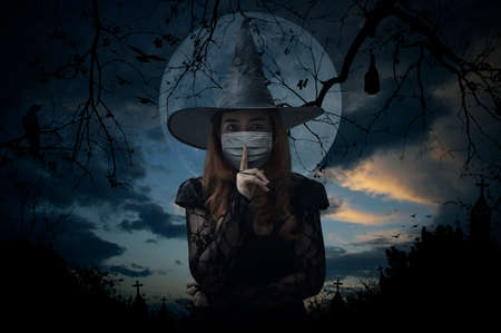 Halloween witch wearing medical face mask showing silence sign with finger over lips standing over cross, church, crow, bat, birds, dead tree, full moon and sunset sky, Halloween conceptの写真素材