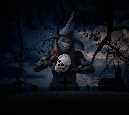 Halloween witch wearing medical face mask holding a skull standing over grass, dead tree, cross, birds with church over spooky cloudy sky, Halloween and coronavirus or covid-19 conceptの写真素材