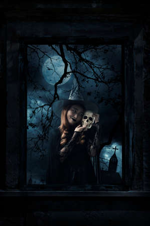 Halloween witch holding a skull standing in old damaged wood window with wall over cross, church,...