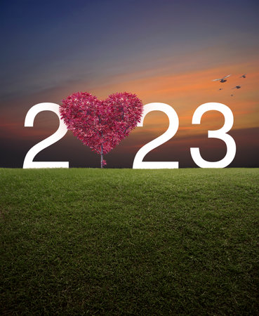 Red tree in the shape of heart love with 2023 white text on green grass field over sunset sky with...