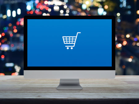 Shopping cart flat icon on desktop modern computer monitor screen on wooden table over blur colorful night light traffic jam road in city, Business shop online conceptの写真素材