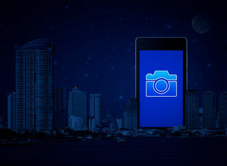 Camera icon on modern smart mobile phone screen over office city tower, river and fantasy night sky, Business camera service shop online conceptの写真素材