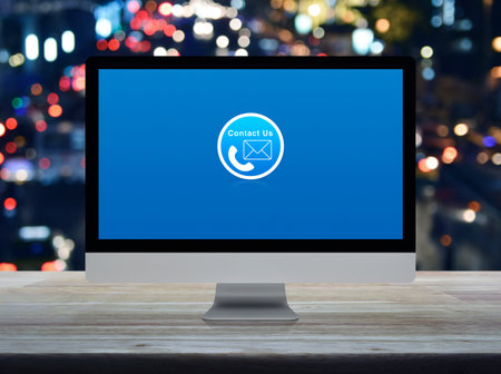Telephone and email flat icon on desktop modern computer monitor screen on wooden table over blur...