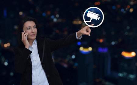 Businesswoman talking on her cell phone and pointing finger to cctv camera icon over blur colorful night light modern city tower and skyscraper, Technology security and safety online conceptの写真素材