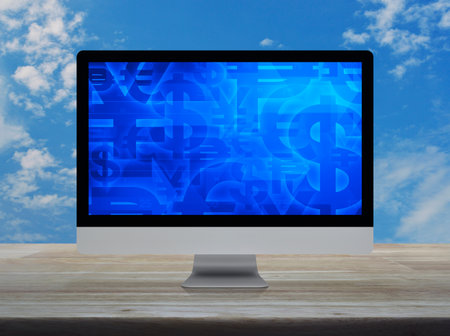 Financial currency symbol on desktop computer monitor screen on wooden table over blue sky with white cloudsの写真素材