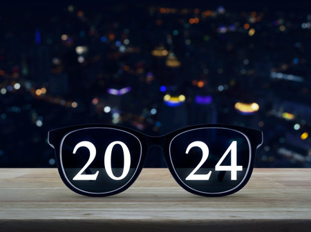 2024 white text with black eye glasses on wooden table over blur colorful night light modern city tower and skyscraper, Business vision happy new year 2024 conceptの写真素材