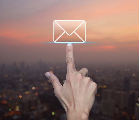 Hand pressing email flat icon over blur of cityscape on warm light sundown, Business contact us online conceptの写真素材
