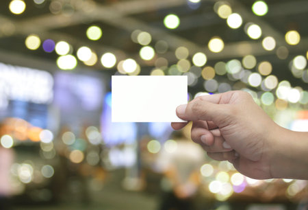 Man hand holding blank white business name card in fingers over blur light and shadow of shopping...