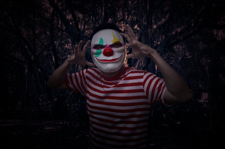 Scary evil clown standing over spooky dark forest with tree, leaves and vine, Halloween mystery conceptの写真素材