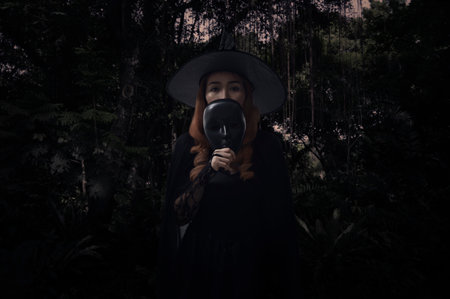 Halloween witch holding black face mask standing over dark forest and tree, Halloween mystery conceptの写真素材