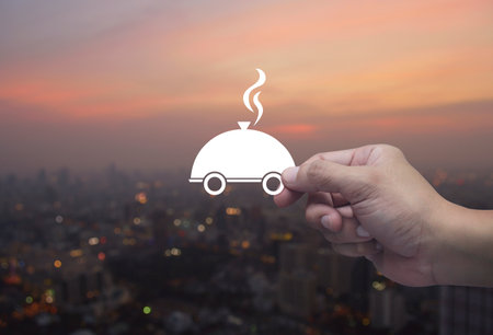 Hand holding restaurant cloche flat icon in fingers over blur of cityscape on warm light sundown, Food delivery online service conceptの写真素材