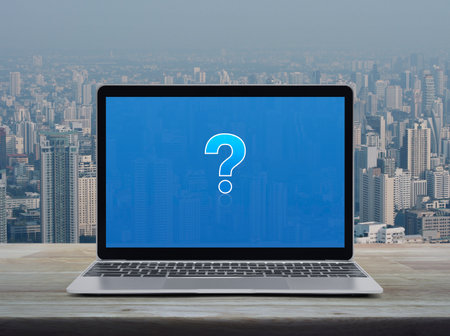 Question mark sign flat icon on modern laptop computer monitor screen on wooden table over city tower and skyscraper, Business customer service and support online conceptの写真素材