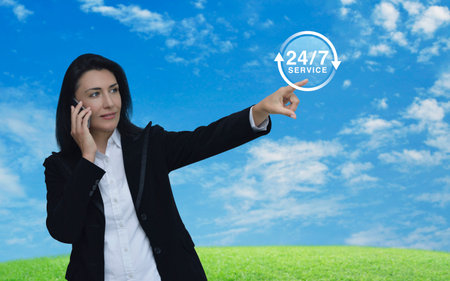 Businesswoman talking on her cell phone and pointing finger to 24 hours service icon over green...