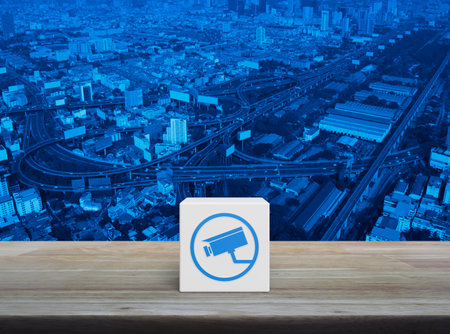 Cctv camera flat icon on white block cube on wooden table over modern city tower, street, expressway...