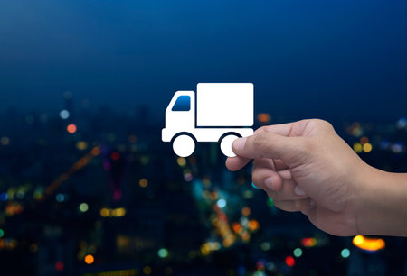 Hand holding delivery truck flat icon in fingers over blur colorful night light modern city tower and skyscraper, Business transportation service conceptの写真素材