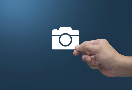 Hand holding camera flat icon in fingers over light blue background, Business camera service shop online conceptの写真素材
