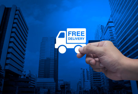 Hand holding free delivery truck flat icon in fingers over modern office city tower and skyscraper, Business transportation service conceptの写真素材