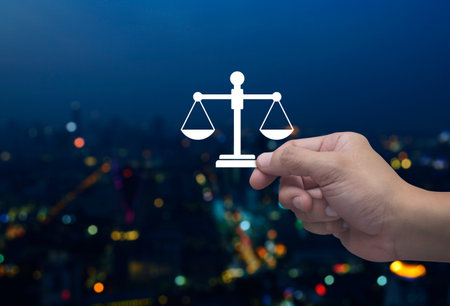 Hand holding law flat icon in fingers over blur colorful night light modern city tower and skyscraper, Business legal service conceptの写真素材