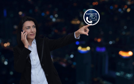 Businesswoman talking on her cell phone and pointing finger to telephone and email icon button over blur colorful night city tower and skyscraper, Business contact us and customer online service conceptの写真素材