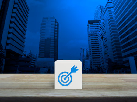 Target with dart flat icon on white block cube on wooden table over modern office city tower and skyscraper, Business success conceptの写真素材