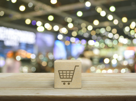 Shop cart flat icon on wood block cube on wooden table over blur light and shadow of shopping mall, Business shopping online conceptの写真素材