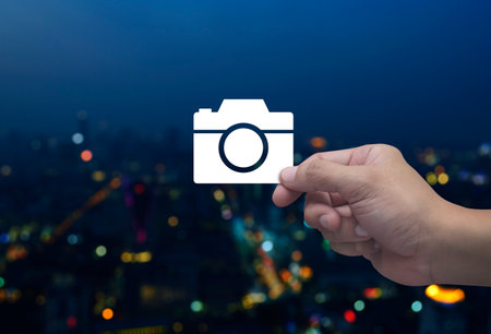 Hand holding camera flat icon in fingers over blur colorful night light modern city tower and...
