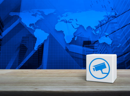 Cctv camera icon on white block cube on wooden table over world map, modern office city tower and...