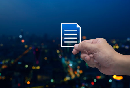 Hand holding document flat icon in fingers over blur colorful night light modern city tower and...