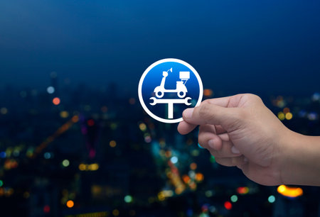 Hand holding service fix motorcycle with wrench tool flat icon in fingers over blur colorful night light modern city tower and skyscraper, Business repair motorbike service conceptの写真素材