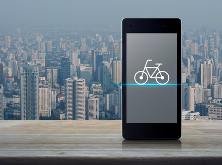 Bicycle flat icon on modern smart phone mobile screen on wooden table over city tower and...