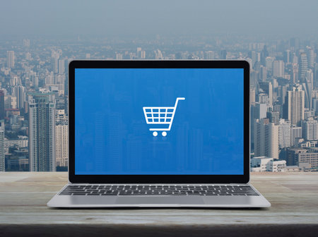 Shopping cart flat icon on modern laptop computer screen on wooden table over city tower and...