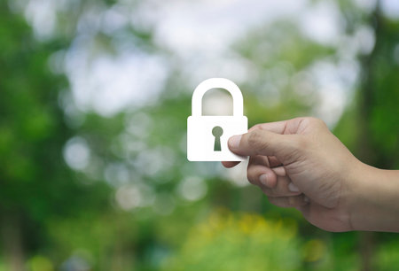 Hand holding padlock flat icon in fingers over blur green tree in park, Technology internet security and safety online conceptの写真素材